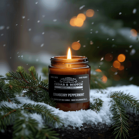 A lit amber glass jar candle labeled Cedar & Scotch Cranberry Peppermint sits outdoors in snow with pine branches around it, against a blurred holiday background with falling snow and warm lights.